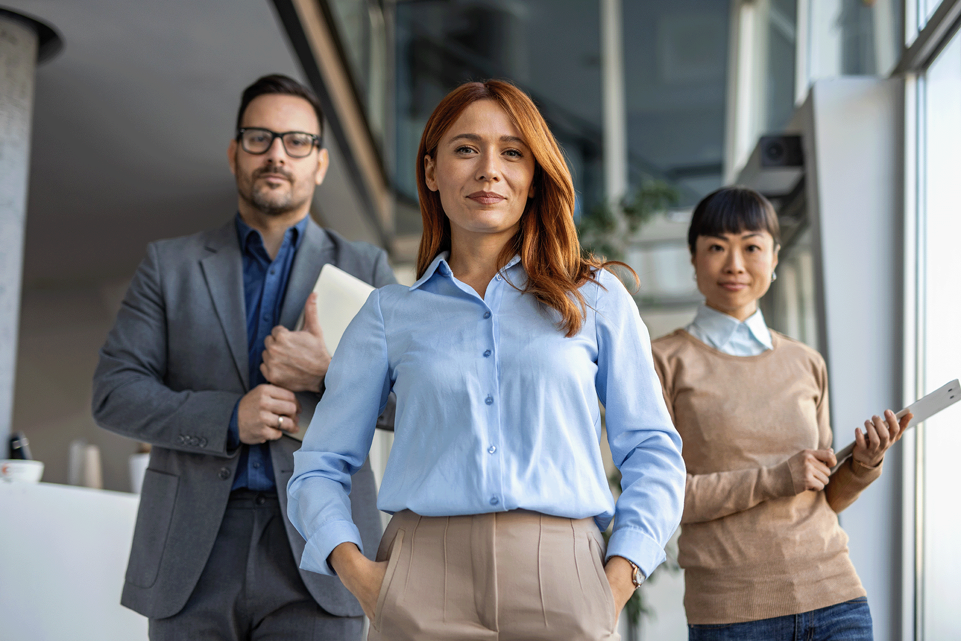 A group of three business people, dressed in business casual attire, holding files, a device, and with hands in their pockets as they look at the camera.