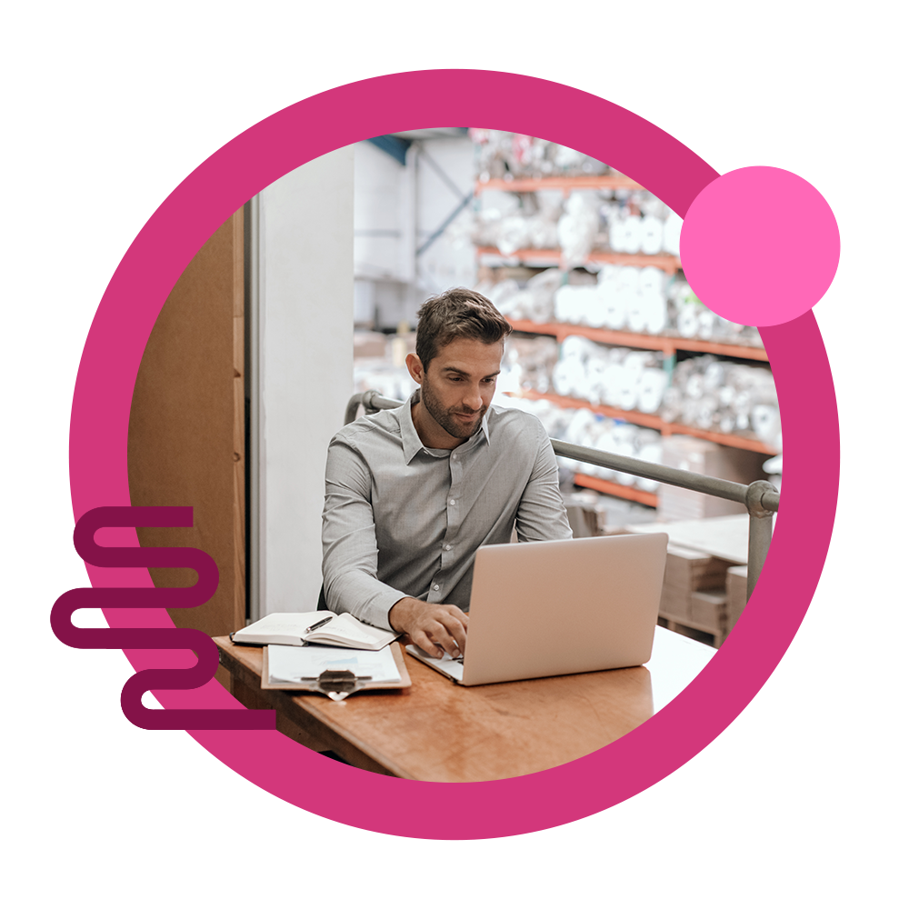 A businessman using his laptop for B2B ecommerce solutions in a warehouse with paper rolls.
