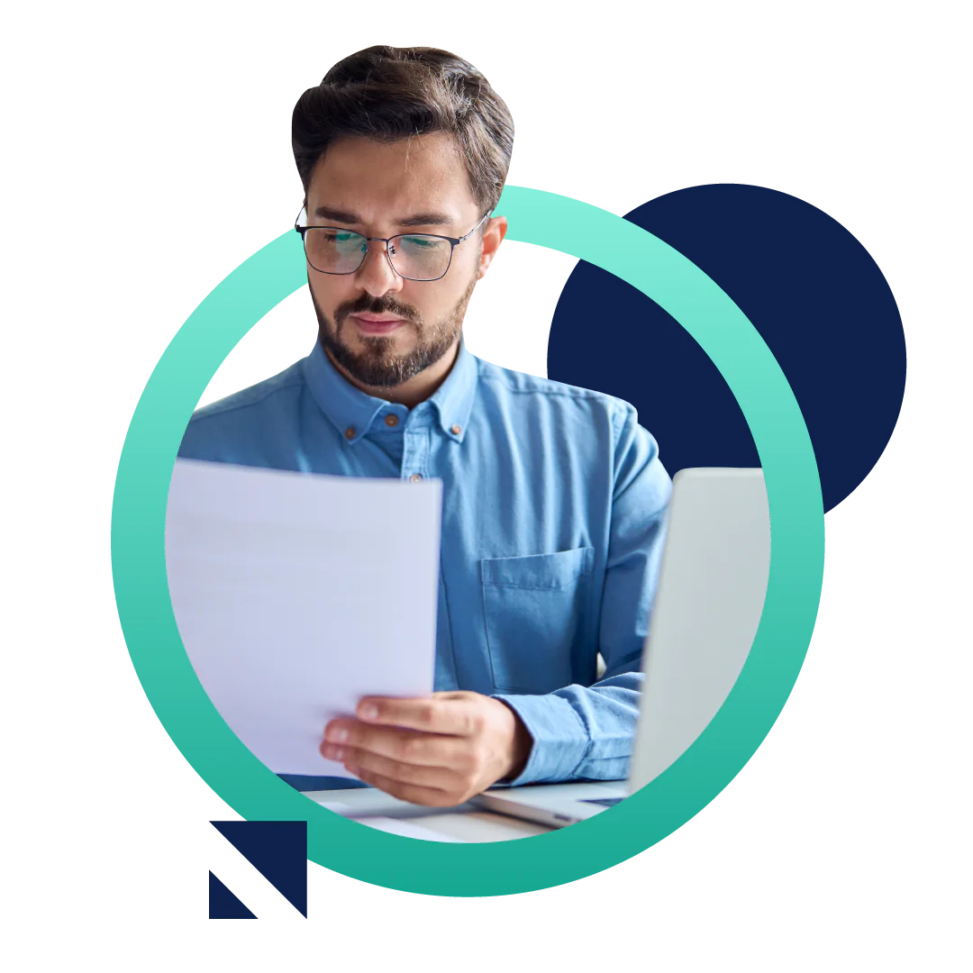 A business man in a blue buttoned shirt looks at a document while sitting at his desk in front of a computer, all placed inside of a designed green circle.