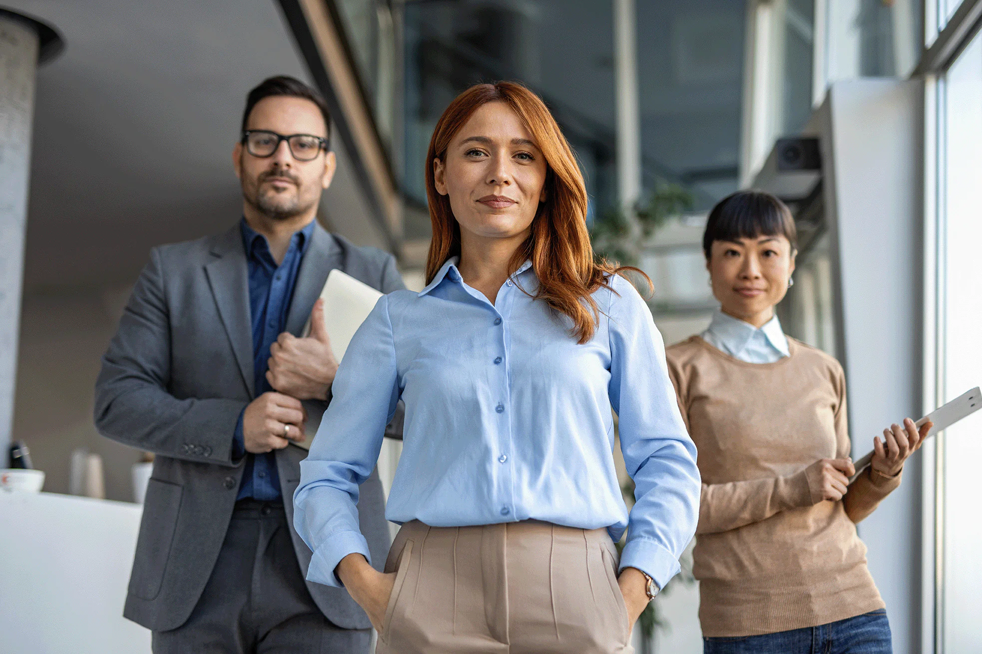 A group of three business people, dressed in business casual attire, holding files, a device, and with hands in their pockets as they look at the camera.