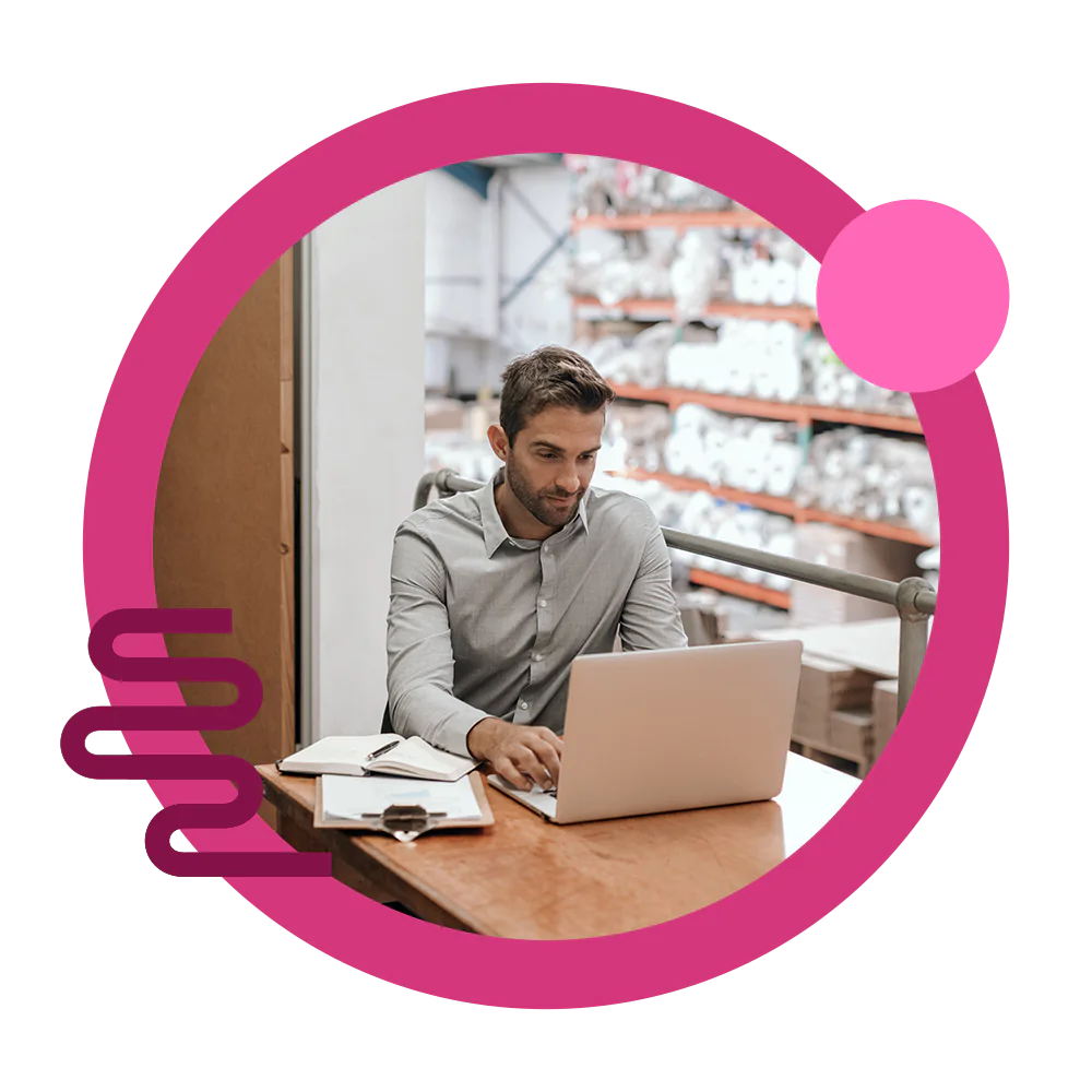 A businessman using his laptop for B2B ecommerce solutions in a warehouse with paper rolls.